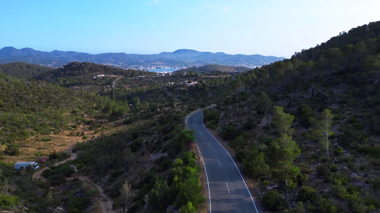 Road winding through a vibrant green forest in Ibiza, Spain, leading down towards the beautiful San Antonio Bay. Unique aerial view flight panorama overview drone