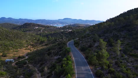 road winding through a vibrant green forest in ibiza, spain, leading down towards the beautiful san antonio bay. unique aerial view flight panorama overview drone