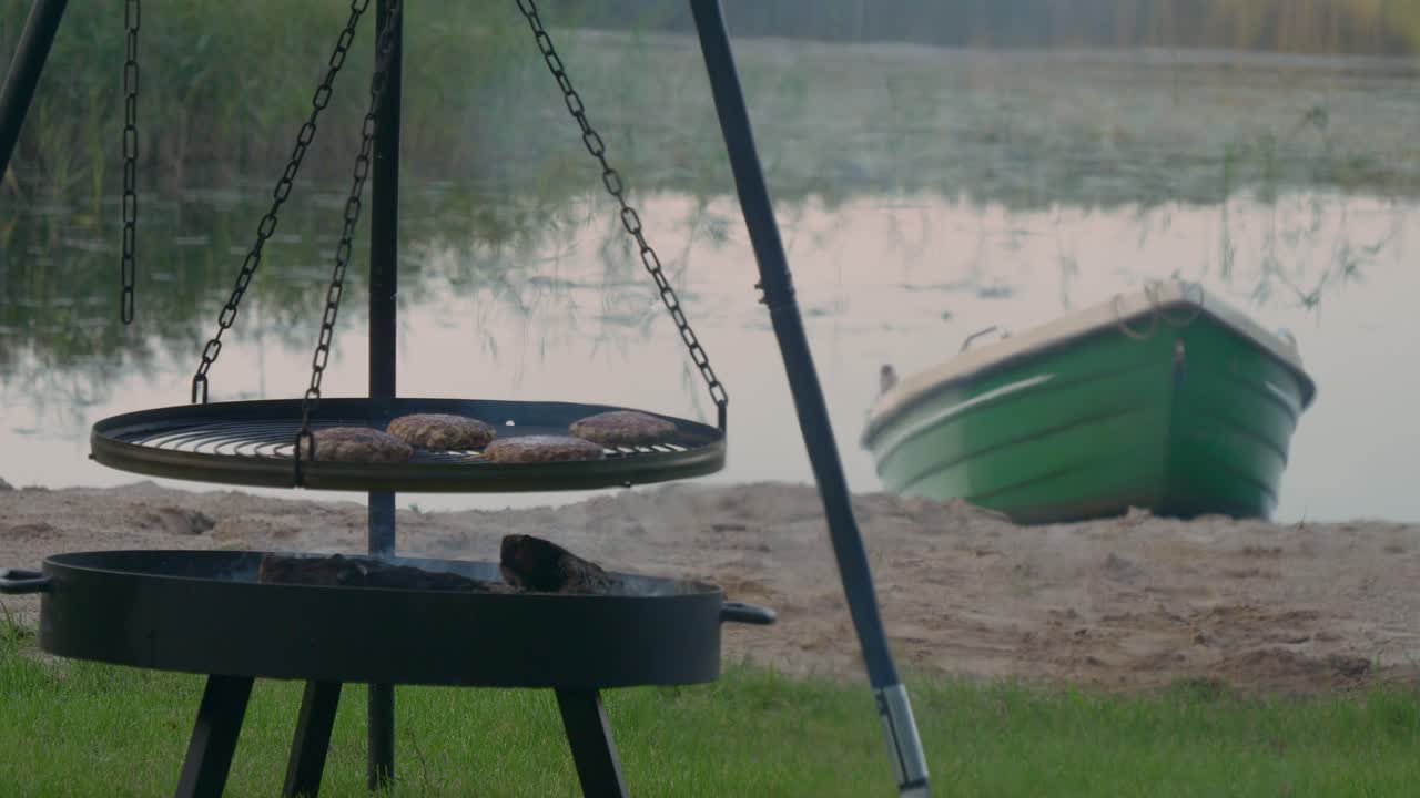Beef Patties Cooked And Grilled On A BBQ Tripod Swivel Hanging Grill At The Lakefront With Wooden Boat In The Background. - wide shot