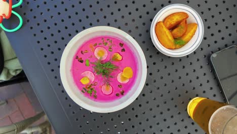 top view of a vibrant pink cold beet soup garnished with herbs and served with a side of potato wedges. a glass of beer and a smartphone are visible on the black table. ideal dining themes