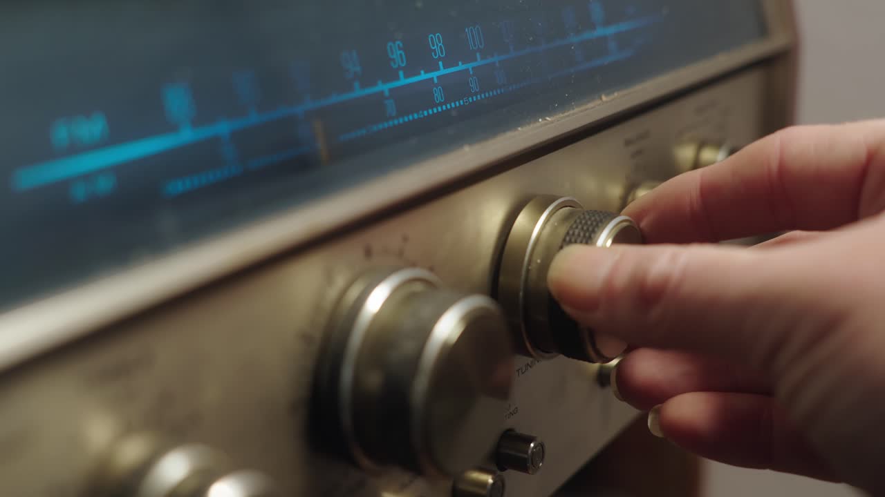 A person&rsquo;s hand , turning knobs to adjust the station on vintage radio with metallic knobs and an analog frequency dial illuminated in blue