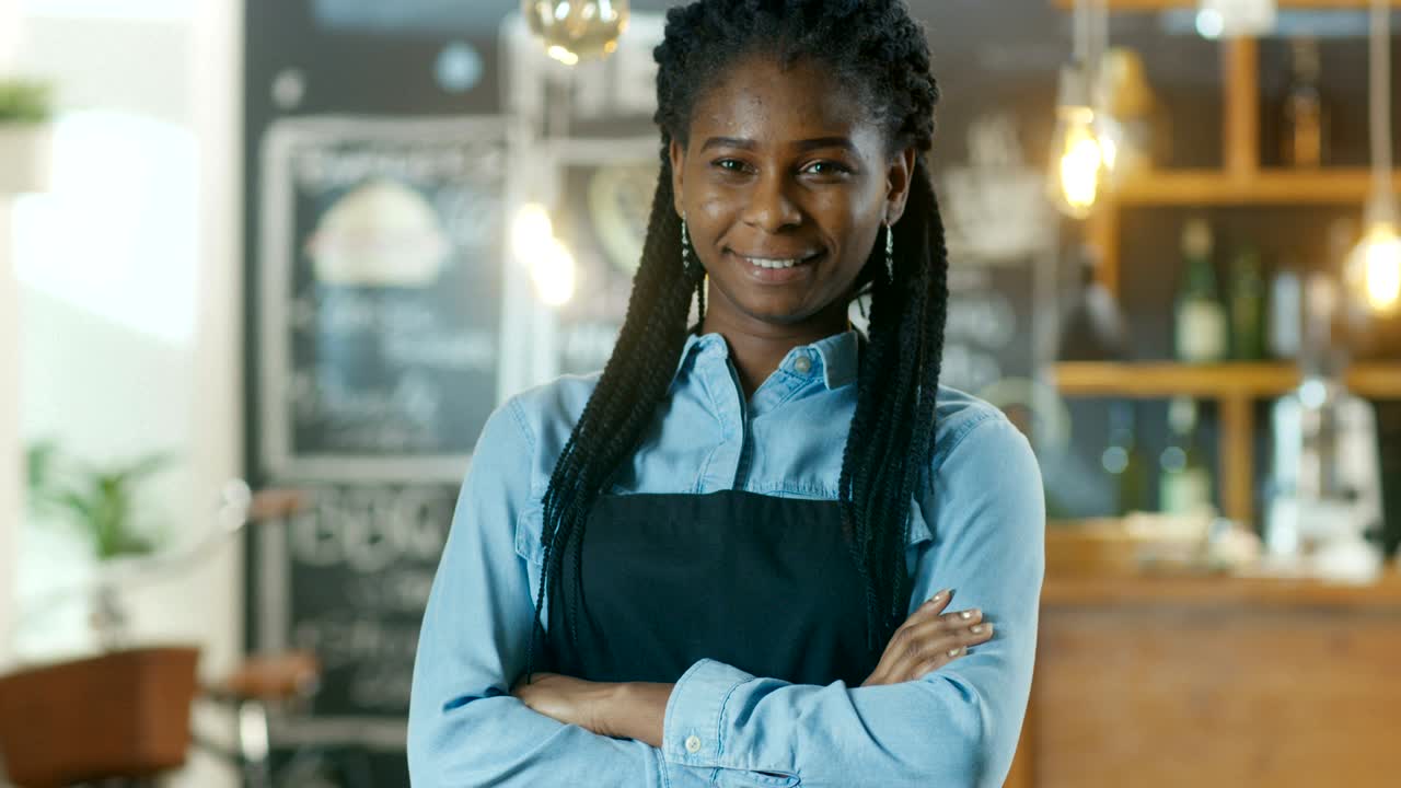 Bella proprietaria di caffè afroamericana in piedi con le braccia incrociate e un sorriso nel suo elegante caffè
