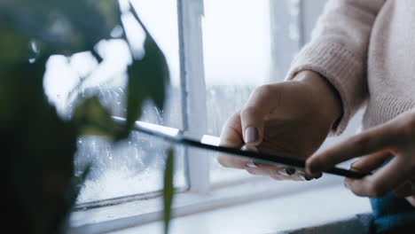 cerca de las manos de la mujer usando una tableta digital navegando por mensajes en línea leyendo redes sociales disfrutando de un dispositivo móvil con pantalla táctil de pie junto a la ventana relajándose en casa en un día frío y lluvioso