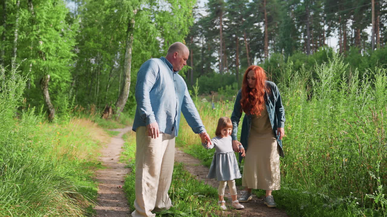 Lekfullt ögonblick mellan litet barn och föräldrar som promenerar på en skogsstig omgiven av gröna träd och solljus, vilket uttrycker värme, samhörighet och glädje under en fridfull familjetid utomhus