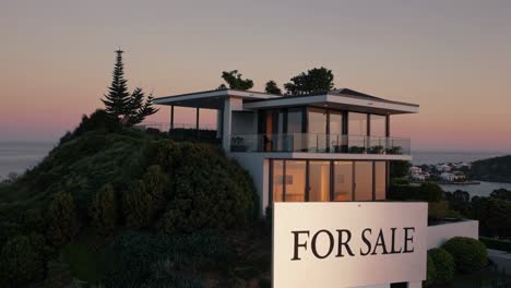contemporary hillside home featuring large glass windows and sleek design, set against a picturesque sunset. surrounded by lush greenery, the property is highlighted by a prominent for sale sign
