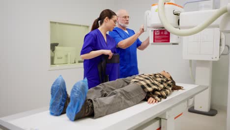 a young brunette girl in a checkered shirt lies on a bed in an x-ray room while experienced doctors an elderly man and a girl in a blue uniform perform x-rays to diagnose a young girl and prevent diseases in a modern clinic