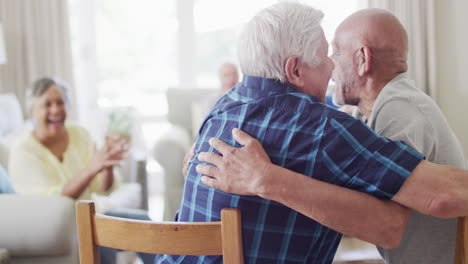 two happy diverse senior men hugging in living room, with diverse senior friends, slow motion