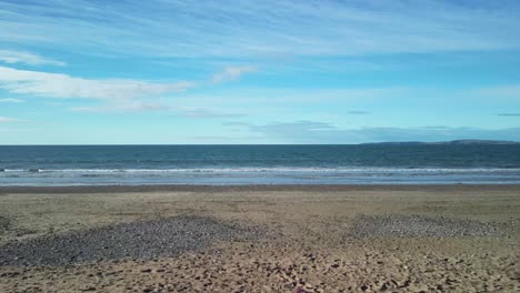 a woman is sitting, a man passing, on a sandy beach while drone takes off over calm atlantic ocean, ireland aerial.