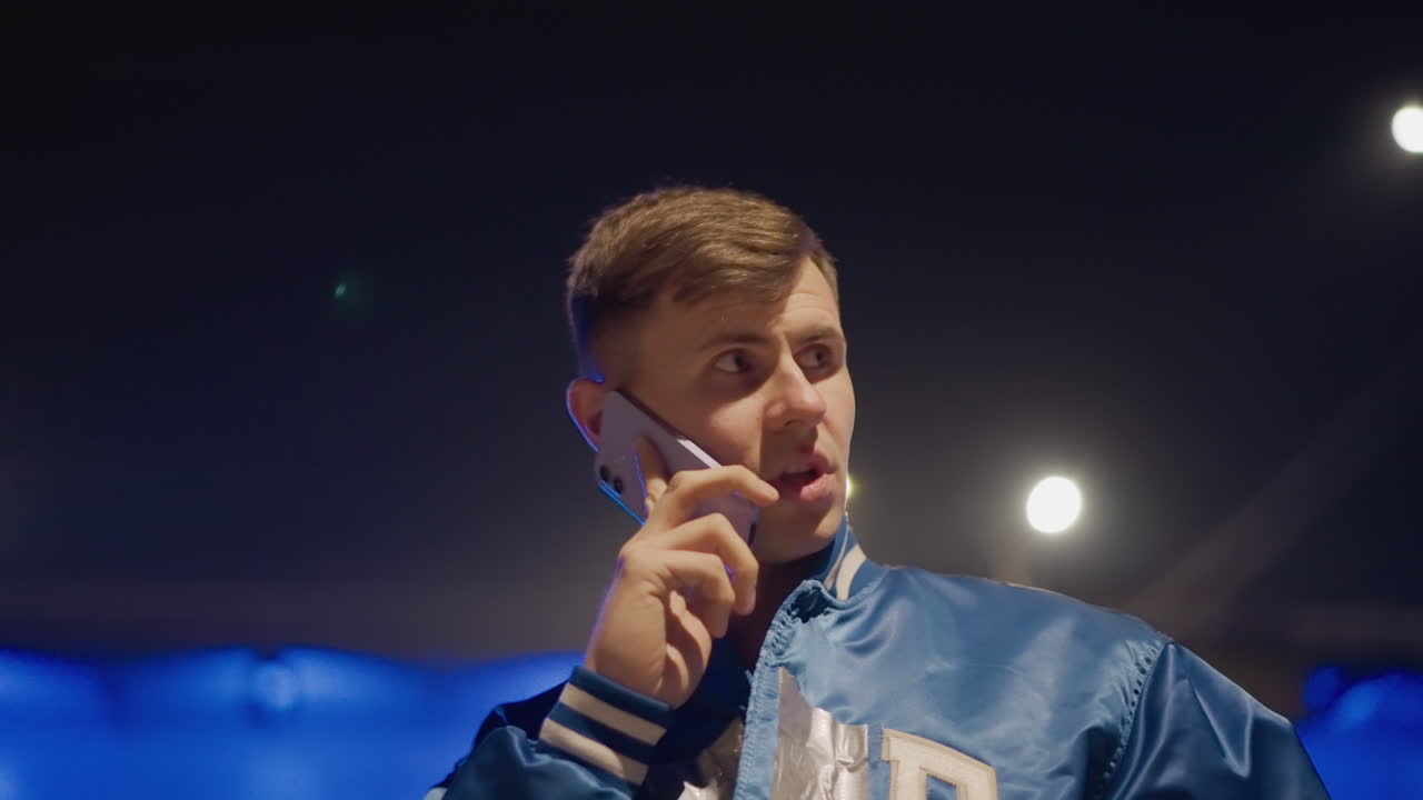 밤거리에서 파란색 새틴 재킷을 입고 휴대폰으로 다급하게 통화하는 젊은 남자의 모습이에요. 보케 가로등 아래, 긴장된 표정으로 고속도로 근처에서 픽업을 기다리고 있어요. 네온 불빛이 반사되는 영화 같은 클로즈업 장면입니다.