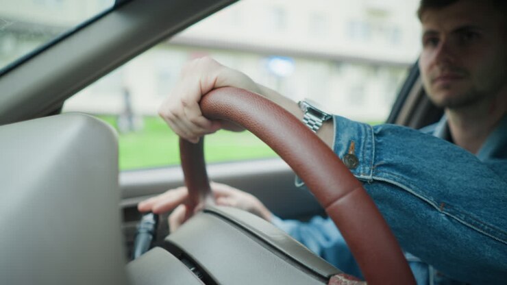 Un homme tenant un volant marron à l’intérieur d’une voiture, bâtiments de banlieue visibles, veste en jean et montre-bracelet, posture détendue, un navetteur faisant une pause à une intersection, vérifiant son rétroviseur et ajustant sa prise en main en douceur