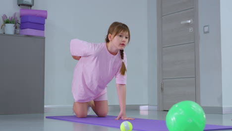 little child on purple mat places pink block on waist while crawling forward during fitness session, with modern gym equipment in background, showing engagement in physical activity