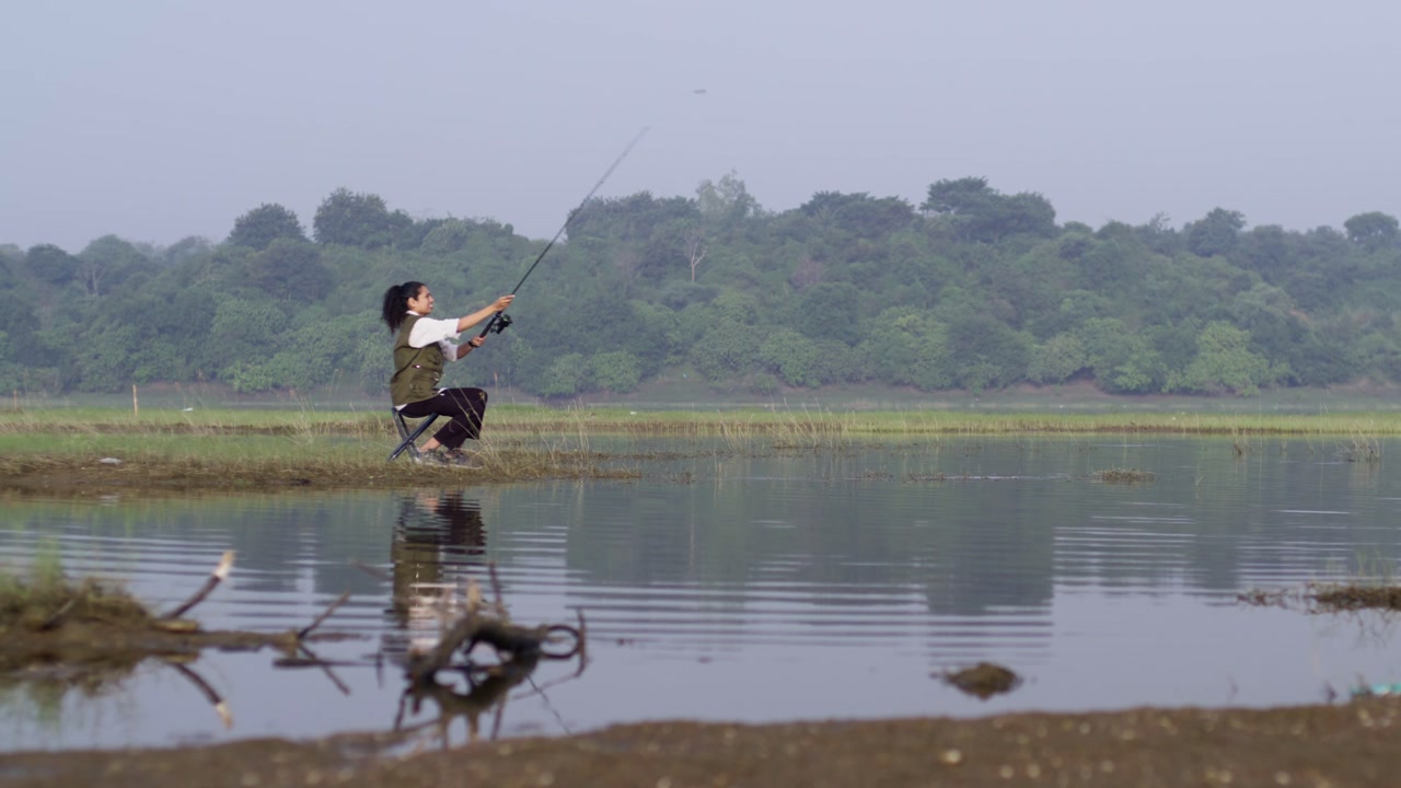 A slow-motion of a South Asian Woman in green vest and black pants, fishing alone on a lush riverbank under misty sky, with forest in the background.