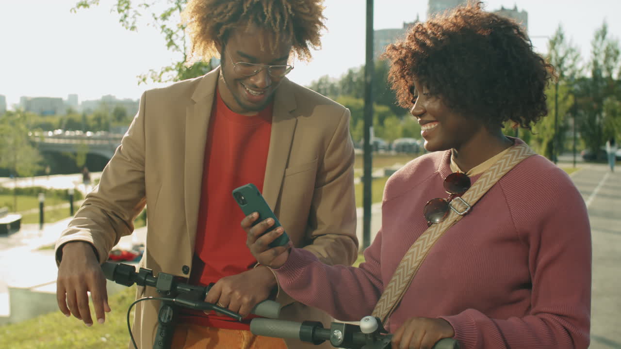 amigos afroamericanos positivos de pie al aire libre con scooters eléctricos y usando el teléfono