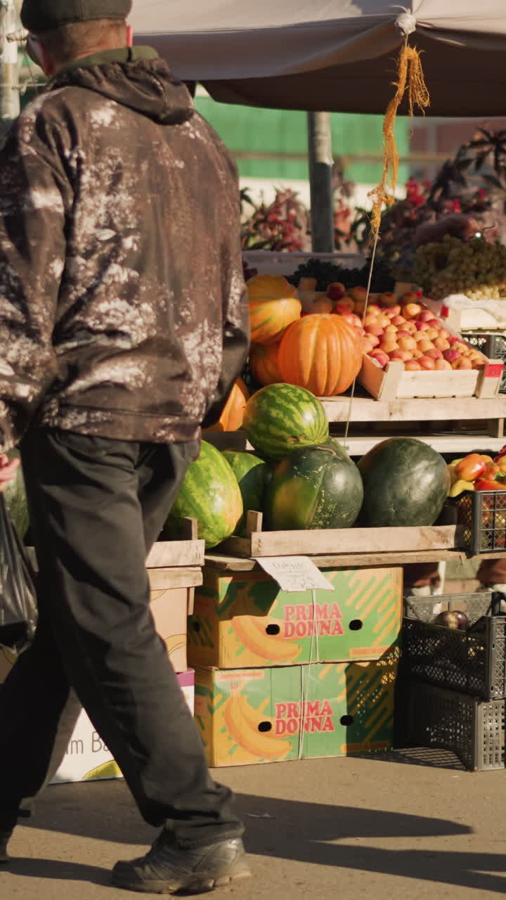 Белый мужчина на фермерском рынке выбирает тыкву и арбуз, несет пластиковый пакет, одет в темную куртку и кепку, осенний солнечный свет создает золотые оттенки, городской уличный прилавок со сложенными ящиками