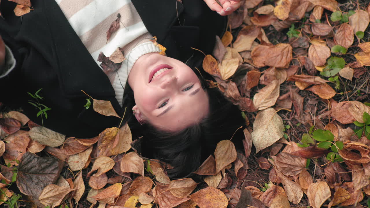 Overhead view of beautiful girl lying face up on autumn forest floor, smiling while blocking dry leaves with hands, cozy sweater and black coat, composition capturing playful peaceful fall vibe