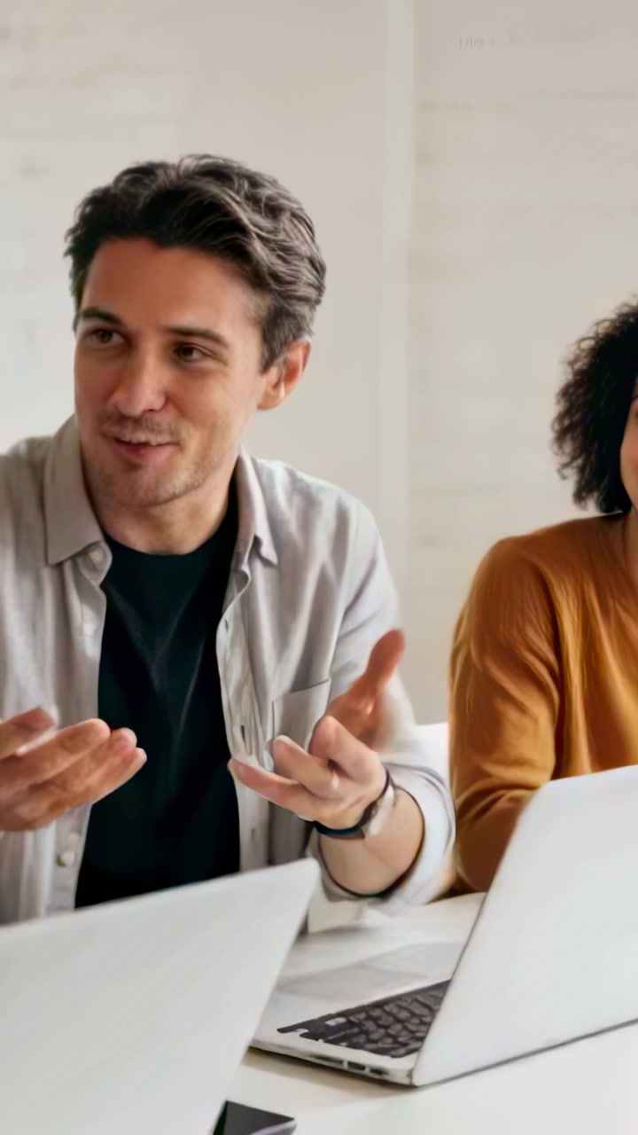 Vertical video: Starting man in gray shirt gesturing and making point at table with laptops phone
