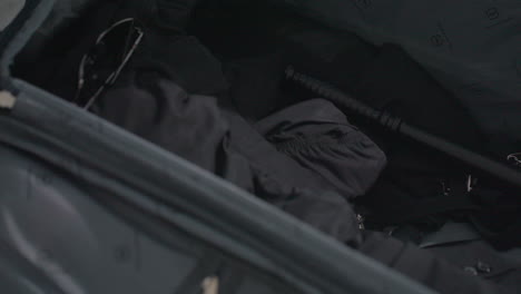 close up of open dark suitcase on floor with hand placing black baton and dark sunglasses inside alongside folded uniform items soft ambient light subtle reflections on metal and fabric textures