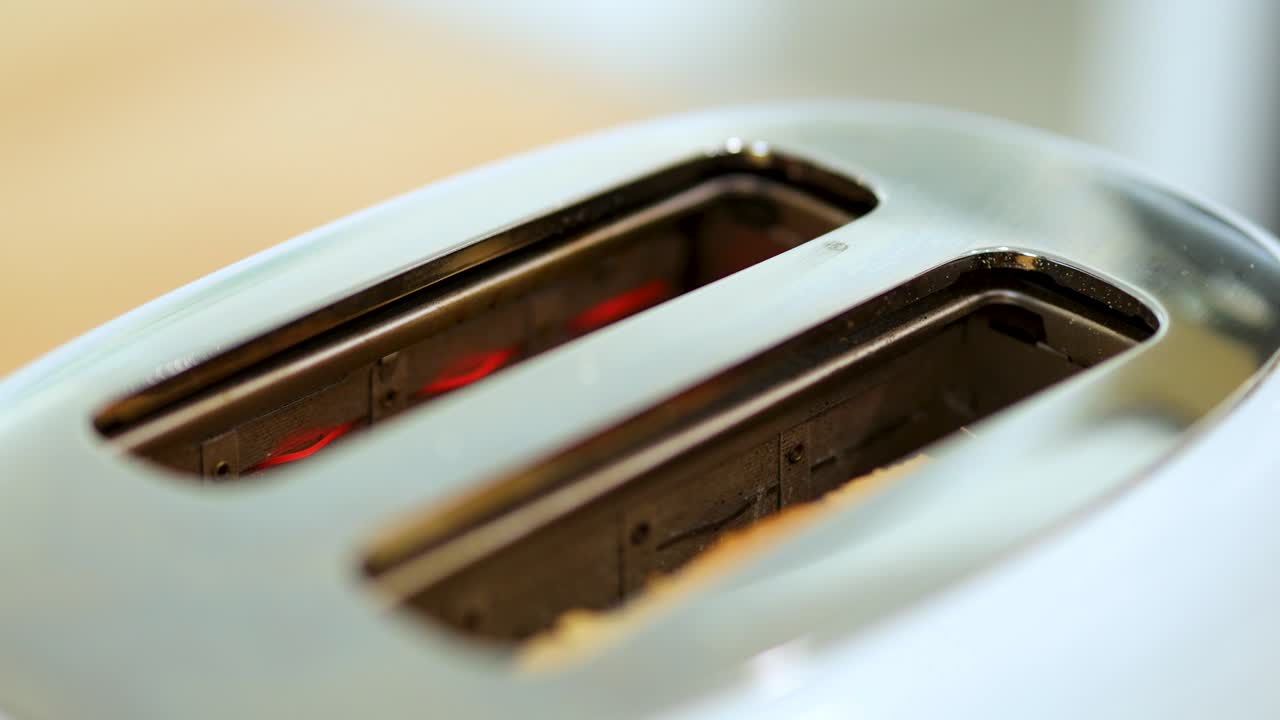 A modern toaster’s slots are shown in close-up as the internal heating elements glow red, indicating active toasting. Soft, natural lighting and shallow depth of field create a warm, domestic atmosphere
