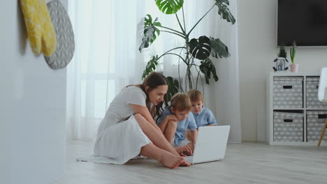 Mamá-Con-Dos-Niños-Pequeños-Sentados-En-El-Suelo-En-Un-Apartamento-Moderno-En-Un-Interior-Luminoso-Con-Una-Computadora-Portátil.-Mire-La-Pantalla-Del-Portátil-Y-Mire-Fotos-Familiares.-Realizar-Compras-Online-Para-Niños.