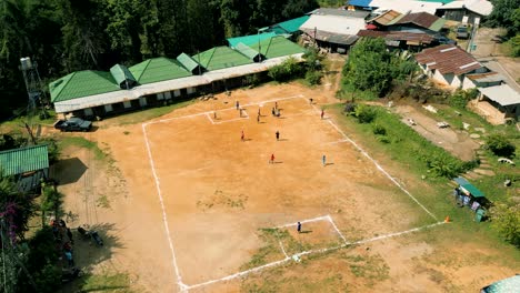 4K-Cinematic-sport-aerial-drone-footage-of-children-playing-on-a-footbal-field-in-the-mountain-village-of-Doi-Pui-next-to-Chiang-Mai,-Thailand-on-a-sunny-day