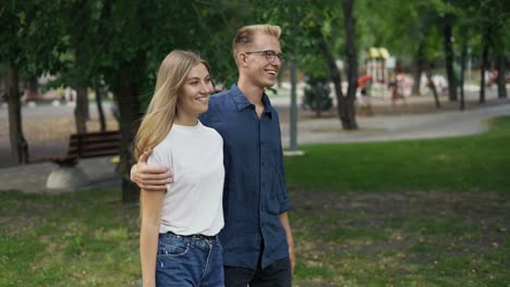 A-young-positive-couple-walks-holding-hands,-meets-in-a-summer-city-park.-The-girl-and-the-boy-are-blonde.-Happy-walk-in-the