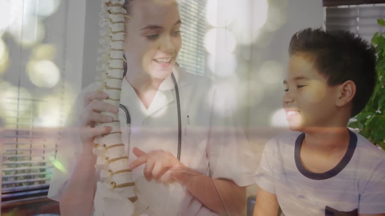 animación digital de puntos brillantes de luz contra una doctora que muestra un modelo de columna vertebral humana a un niño
