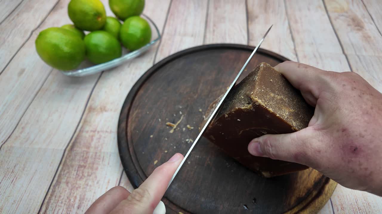 Primer plano de unas manos usando un cuchillo para cortar un bloque de panela sobre una tabla rústica de madera, listo para una dulce receta de limonada de caña de azúcar