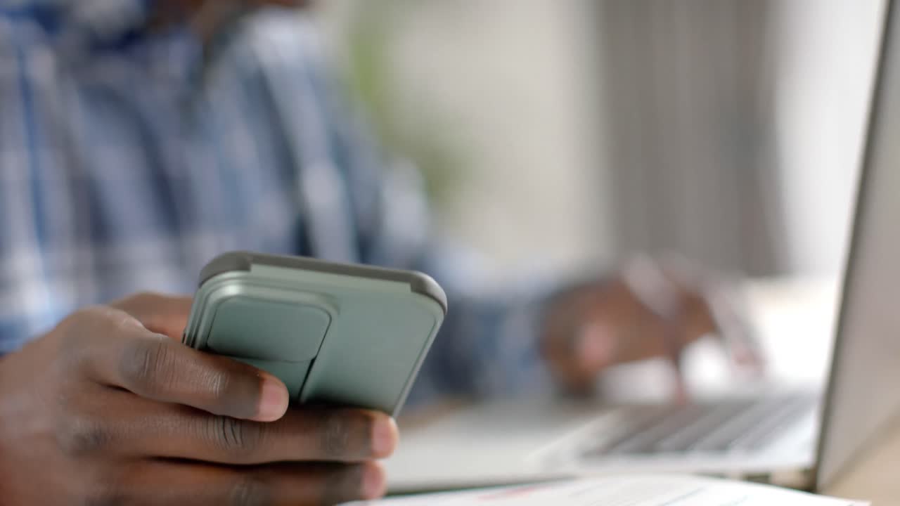 Midsection of african american man at table using laptop and smartphone at home, slow motion