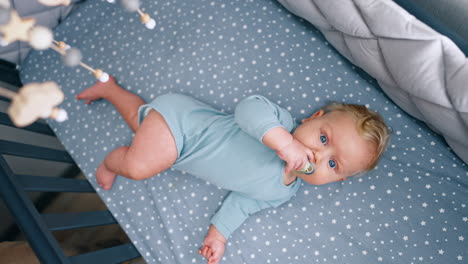 adorable little baby boy in blue bodysuit lies in the crib. cute child with pacifier in mouth looks at cot mobile. top view.