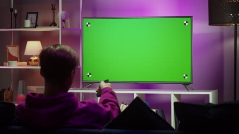 man watching television in living room in evening. young guy sitting on sofa against tv with chroma green screen, back view. male person switching channels using remote controller.