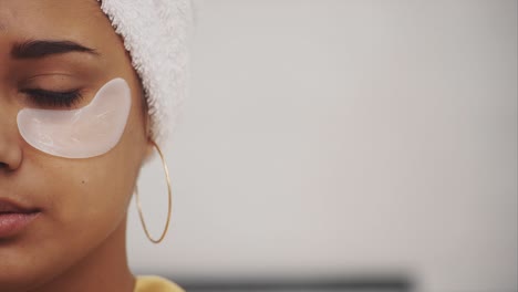 closeup portrait of beautiful woman after bath with towel on head puts patches under the eyes from wrinkles and dark circles. attractive female in bathrobe makes beauty treatments on the face at home.