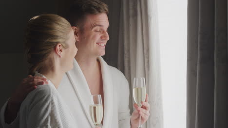 couple wearing bathrobes on romantic hotel break standing by open curtains with glasses of champagne