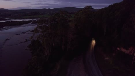 Car-travels-with-headlights-along-dark-moody-unlit-coastal-road-AERIAL