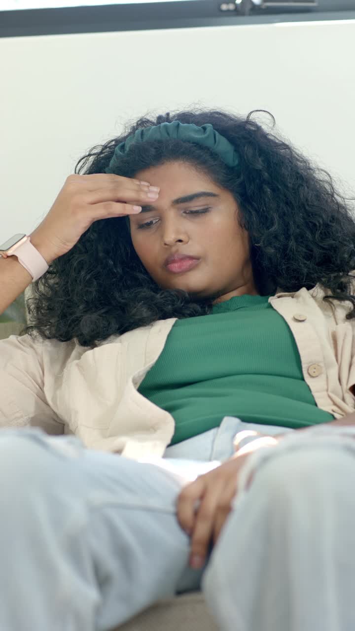 Vertical video: Woman settling back on couch, rubbing forehead, lowering gaze, showing fatigue