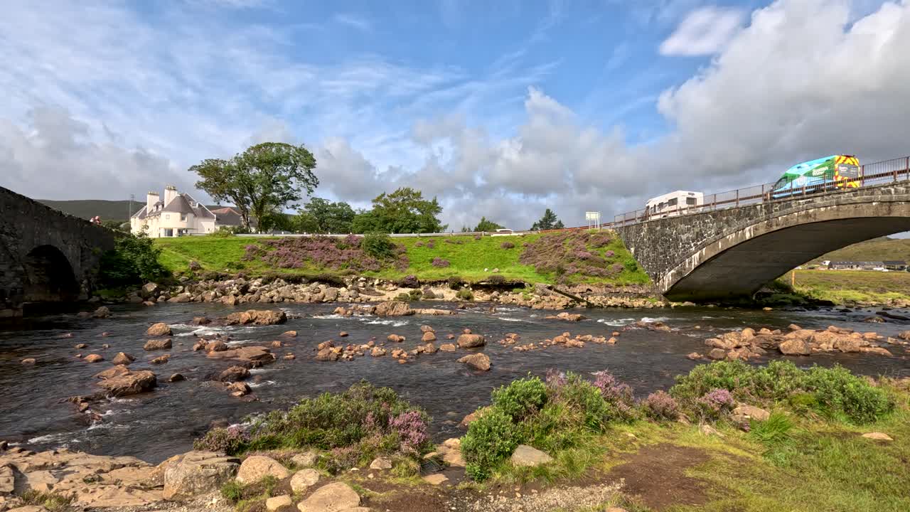 A white camper van drives across a stone bridge above a rocky stream in rural Isle of Skye, under bright daylight with scattered clouds