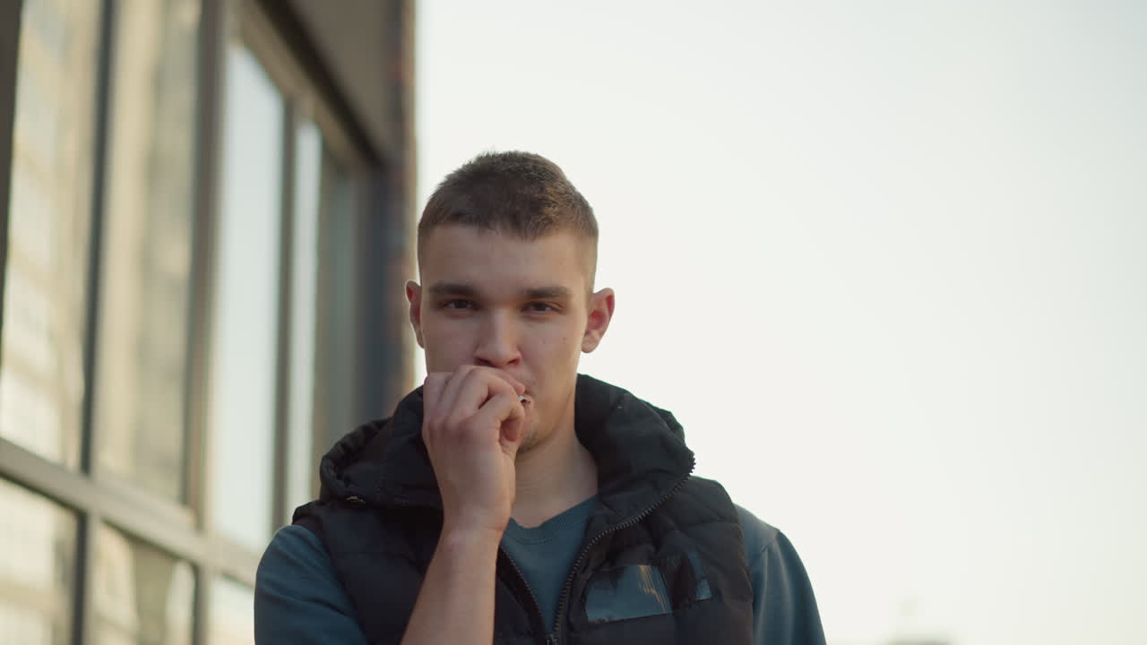 young man in casual outfit walks toward camera holding cigarette in mouth then flicks it away near modern building exterior with soft lighting and thoughtful expression