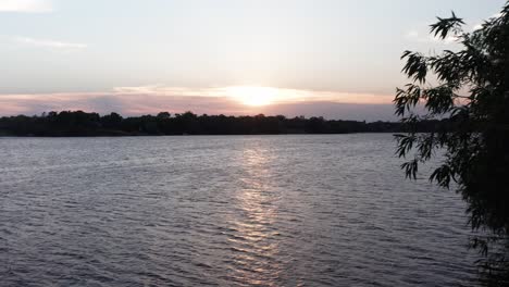 Low-and-slow-aerial-shot-gliding-past-tree-across-open-lake-at-sunset