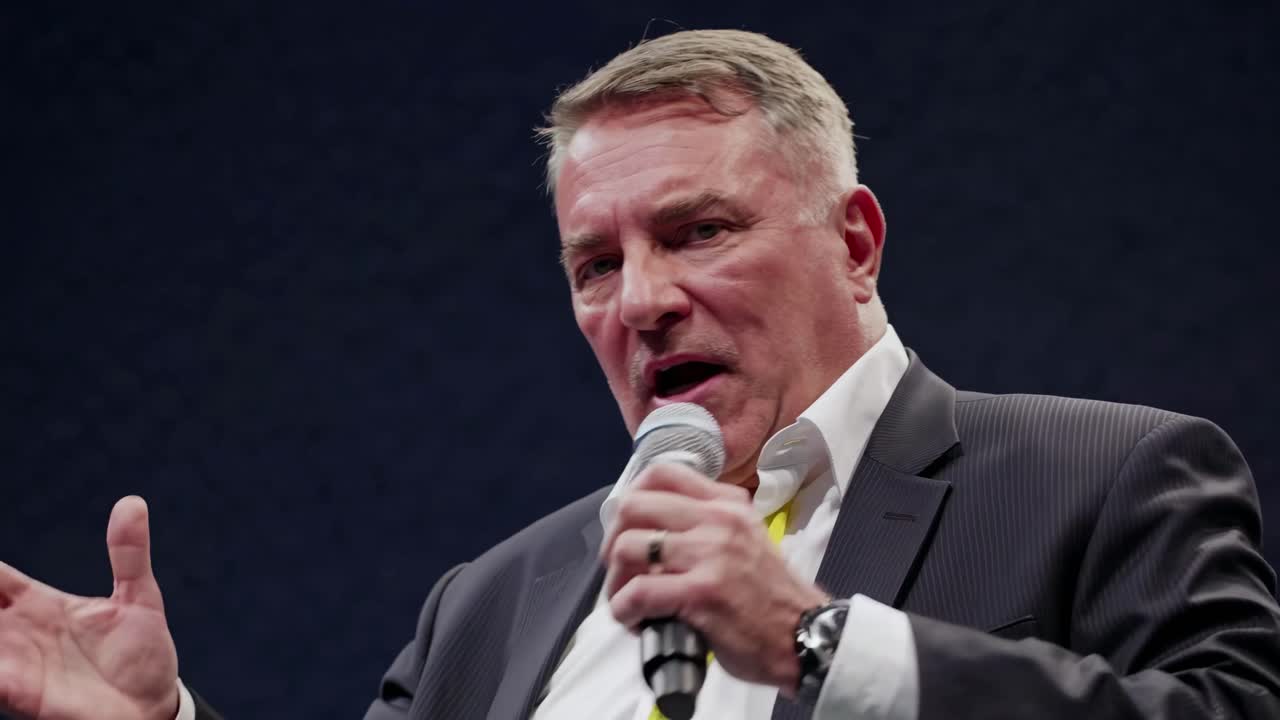 A mid-shot video captures a confident speaker in a suit, gesturing with a microphone against a dark