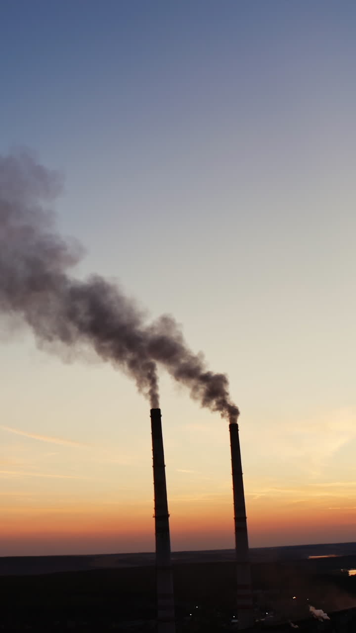 Factory with pipes among nature in the evening. Two industrial pipes with dark smoke pollute the environment at sunset. Vertical video