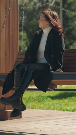 lady in black coat and leather boots sitting on wooden swing chair in park, hair swaying with wind, bag resting on wooden pillar, surrounded by lush greenery