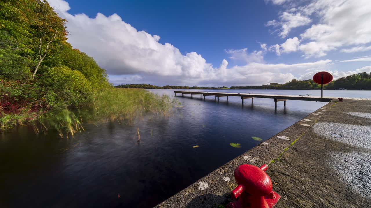 Time-lapse di un molo in legno sul lago in primo piano e un'isola con rovine di castello e foresta in lontananza, in una giornata nuvolosa e soleggiata a Lough Key nella contea di Roscommon in Irlanda.