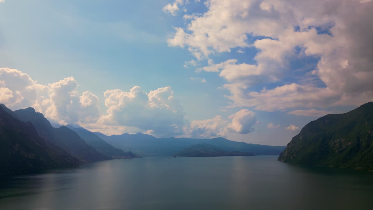 Expansive aerial view sweeps across placid Lake Iseo toward distant Lombardy mountains under billowing summer clouds, conveying serenity and scale. Lake Iseo, Lago d'Iseo