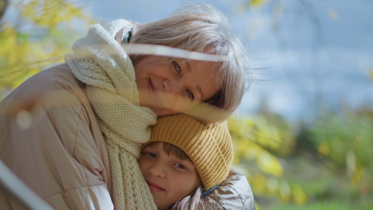 mother hugs daughter warmly under canopy of golden autumn leaves showing joy and tender bond, both bundled in colorful coats and soft scarves, with sunlight filtering through trees in serene