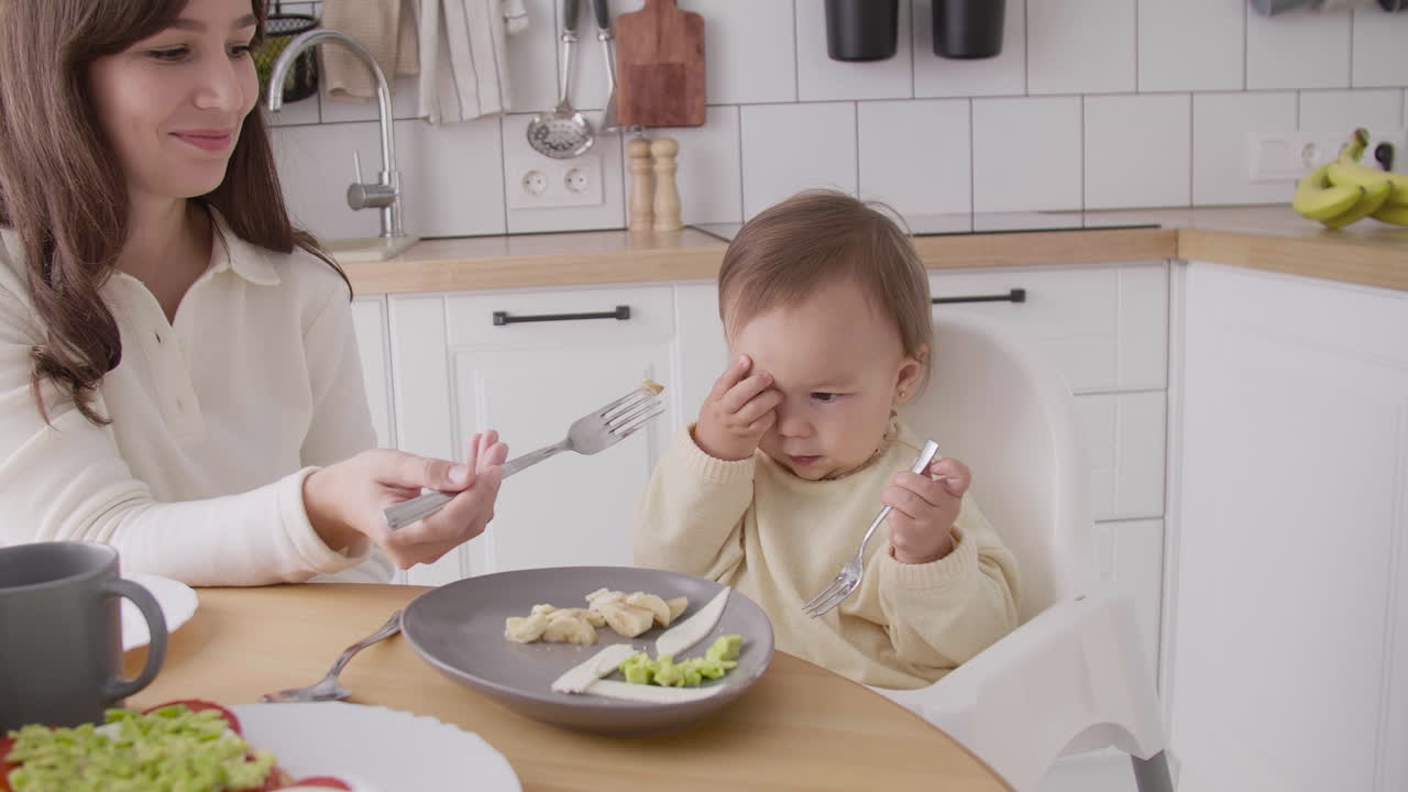 Mor fodrer sin uvillige lille pige, der sidder i højstol i køkkenet.