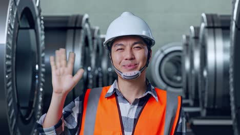 close up of asian male engineer with safety helmet smiling to camera and waving hand saying bye while standing in metal factory