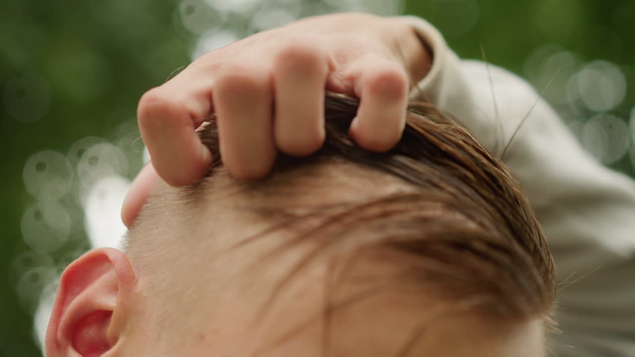 젊은 남자가 가벼운 비가 온 후 젖은 머리를 손가락으로 스타일링하는 모습, 잎이 무성한 배경, 부드러운 보케, 자연광, 사색적인 분위기, 라이프스타일 영상 클립, 흐린 하늘 아래 야외에서 젖은 머리 다듬기 클로즈업