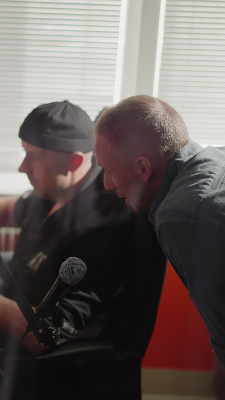 Two men are in a studio room, one seated in front of a computer, likely a producer, wearing black clothes and a cap, while the other, dressed in a long sleeve shirt, leans over, explaining