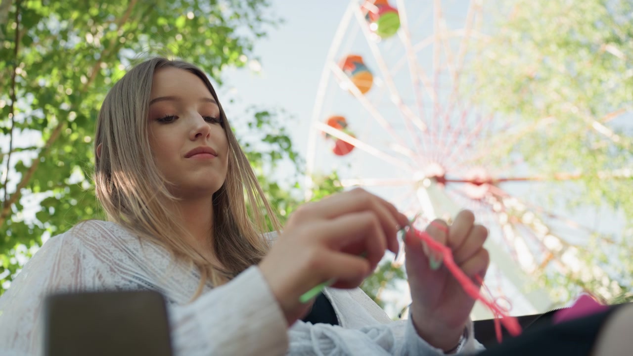 Primo piano di una giovane donna che lavora all'uncinetto con filato rosa brillante in un parco all'aperto, tenendo un uncinetto verde. Lo sfondo presenta alberi rigogliosi e una grande ruota panoramica sotto un cielo sereno.
