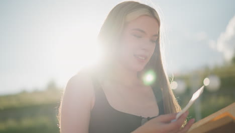 woman seated outdoors holding book finds hidden paper between pages, looking at it with excitement and curiosity under bright sunlight, background shows soft greenery and a blurred lamp post