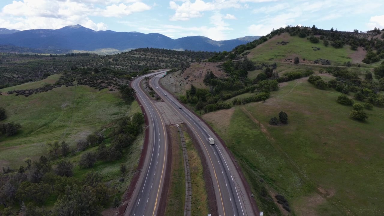 Autostrada a più corsie della California con traffico leggero di camion che si snoda attraverso dolci colline verdeggianti e lussureggianti, alberi sparsi e una catena montuosa innevata sotto un cielo azzurro. California, USA. Ampia ripresa aerea in ascesa